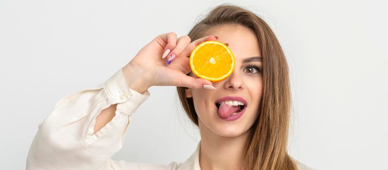 Portrait of a cheerful caucasian young woman covering eye with an orange slice and stick out tongue wears white shirt against a white background