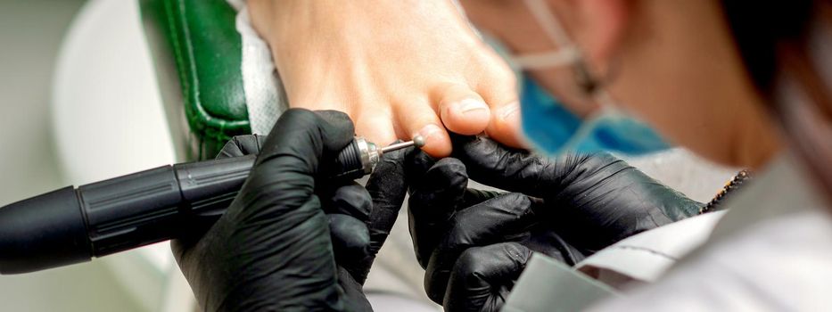 Pedicure master removes corns and cuticles with electric file machine on female toes in a nail salon
