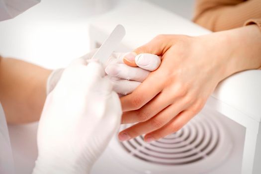 Manicure master wears white gloves with nail file doing manicure on female fingernails in a nail salon