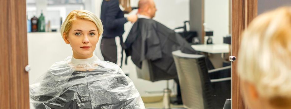 Young female client waiting for the hairdresser looking in the mirror in a hair salon, selective focus