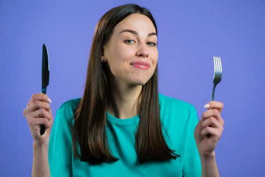 Portrait of hungry woman with fork and knife. Lady waiting for serving dinner dishes with cutlery on violet studio background. High quality photo