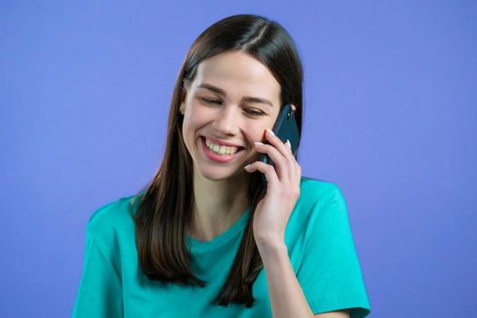 Young woman speaks with smile on phone. Girl holding and using smart phone. Violet studio background. High quality photo