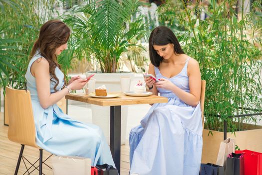 Two young caucasian women sitting at the table and looking at the smartphones with shopping bags in a cafe outdoors