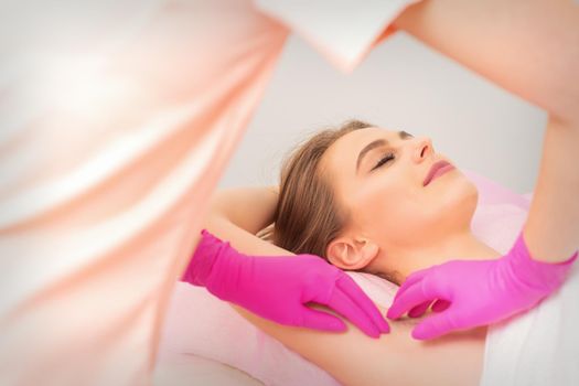 Young caucasian woman receiving waxing armpit by hands in gloves of beautician in hair removal salon