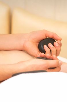Hands of masseur hold black hot stones massaging back of a young adult woman in spa salon