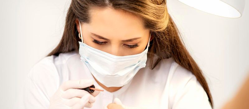 Manicure master in the white protective mask is painting nails with nail polish in a nail salon