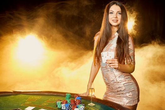 Gorgeous young woman in evening dress with two cards in hands standing near poker table with glass of champagne