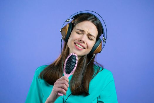 European woman singing and dancing with hair brush or comb instead microphone at purple studio background. Lady in headphones having fun, listening to music, dreams of being celebrity. High quality