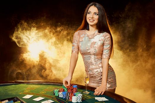Gorgeous young woman in evening dress with chips in hand standing near poker table with glass of champagne