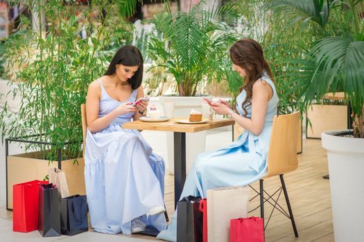 Two young caucasian women sitting at the table and looking at the smartphones with shopping bags in a cafe outdoors