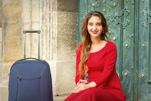 Portrait of a beautiful young caucasian traveling woman sitting at the door with suitcase smiling and looking at the camera outdoors