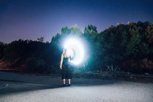 Long exposure of female drawing a portal shaped spiral with a bright light source before a starry night at a deserted rural scenery.