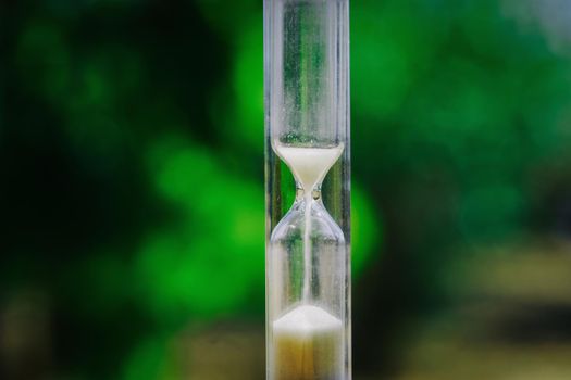 Transparent vertical hourglass with time passing falling sand against green bokeh background.