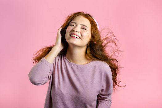 Attractive woman with red hair with headphones on pink studio background. Cute girl portrait. Music, radio, happiness, freedom, youth concept