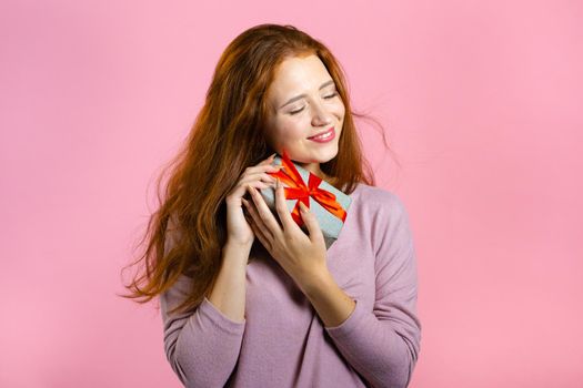 Excited woman received gift box with bow. She is happy and flattered by attention. Girl smiling with present on pink background. Studio portrait.