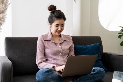 Smiling asian young woman working on laptop at home office. Young asian student using computer remote studying, virtual training, e-learning, watching online education webinar at house.