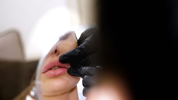 medical office, beautiful woman looking in the mirror, doctor probes the patient's lips after injections of hyaluronic acid into lips, correction of the shape of the lips. High quality photo