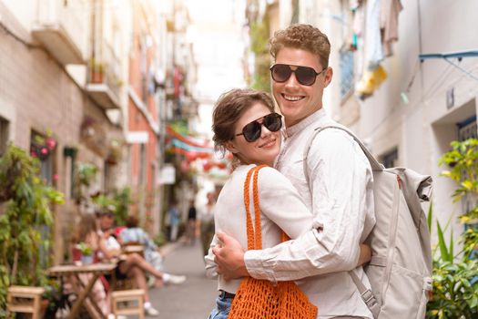 Young beautiful hipster couple in love walking on old city street with shopper and backpack, summer Europe vacation, travel, fun, happy, smiling, sunglasses, trendy outfit, romance, date, embracing