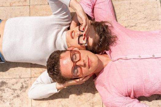 Portrait of two attractive students man and woman wearing stylish glasses lying on floor outdoor and smiling, head to head cheek to cheek, break relax between lesson, work, projects Eyeglasses concept