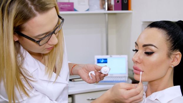 medical office, doctor puts on patient lips a cosmetic cream with a cotton swab, after injections of hyaluronic acid into lips, cleans the surface with an antiseptic. High quality photo