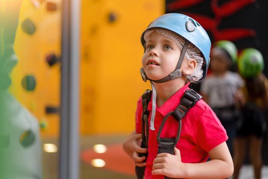 Cute little girl climber in blue protective helmet and gear for climbing standing in climber centre amusement park for children. Caucasian female climber 5 years