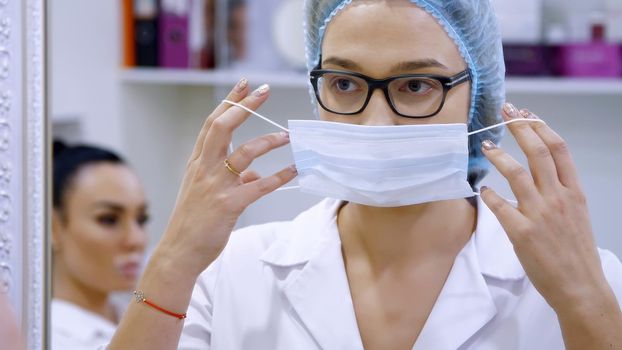 cosmetology room in the clinic, a female doctor wearing glasses, wearing a white uniform, dresses a medical cap and bandage. stands in front of a mirror. High quality photo