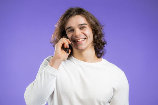 Young smiling man speaks on phone. Guy holding and using smart phone. Violet studio background. High quality photo