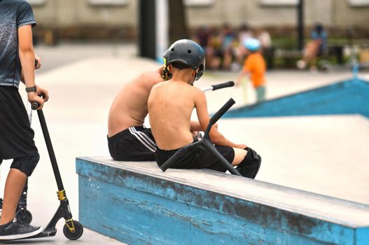 two guys without a t-shirt sit on the training ground for skateboarders. teenage boy with scooters in skate park. Public playground for a skateboard in a recreation park. Sports equipment for kids