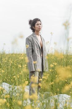 An Asian model poses in a field of yellow flowers for a clothing brand, polyethylene is the main props for a photo shoot. The concept of manufacturing clothing from recycled plastic. A woman in a pantsuit is standing on a plastic bag.