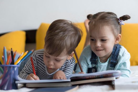 smart smiling preshool toddler children boy and girl draw with pencils lying on their stomach at yellow bed. little cute reader have fun, happy kid on quarantine at home. Friendship, family, education.