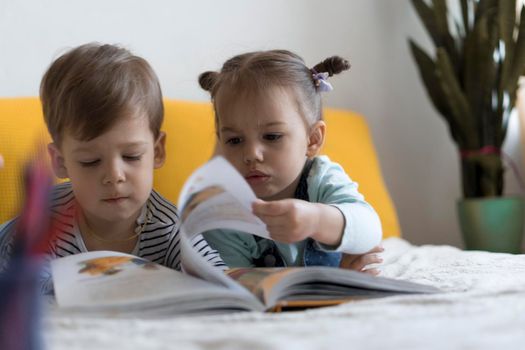 Two preshool, toddler children read large interesting book of fairy tales on yellow bed. Siblings little twins reader have fun, happy kid on quarantine at home. Friendship, family, education concept.