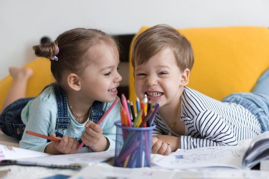 smart smiling preshool toddler children boy and girl draw with pencils lying on their stomach at yellow bed. little cute reader have fun, happy kid on quarantine at home. Friendship, family, education.