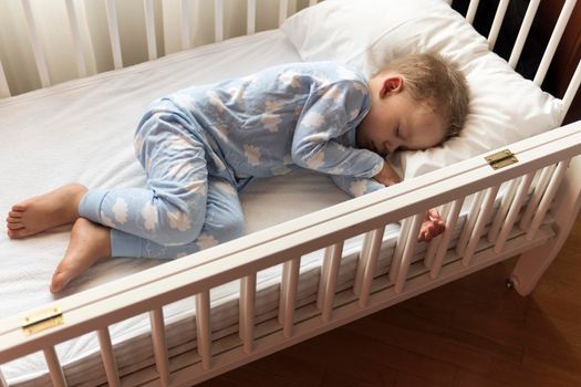 top view Cute little 2-3 years preschool baby boyl kid sleeping sweetly in white crib during lunch rest time in blue pajama with pillow at home. Childhood, leisure, comfort, medicine, health concept.