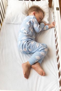 top view Cute little 2-3 years preschool baby boyl kid sleeping sweetly in white crib during lunch rest time in blue pajama with pillow at home. Childhood, leisure, comfort, medicine, health concept.