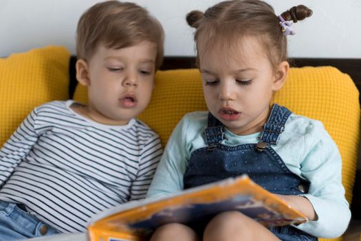 Two preshool, toddler children read large interesting book of fairy tales on yellow bed. Siblings little twins reader have fun, happy kid on quarantine at home. Friendship, family, education concept.