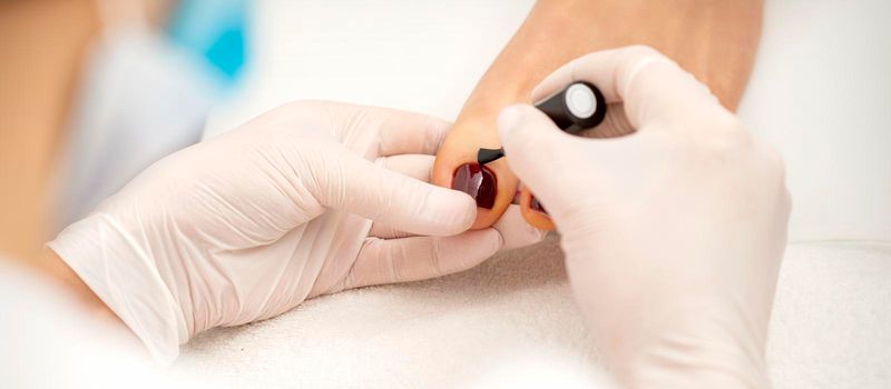Closeup of hands in white gloves applying burgundy nail polish with a brush on woman's toes in a nail salon