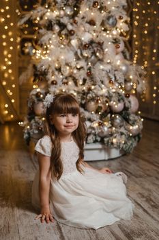 A girl in a festive light dress with a gift in her hands next to a Christmas tree, lights of garlands in the background. The concept of New Year holidays.