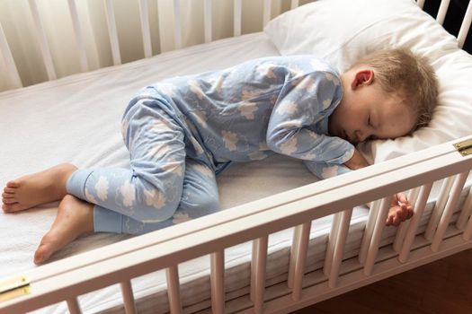 top view Cute little 2-3 years preschool baby boyl kid sleeping sweetly in white crib during lunch rest time in blue pajama with pillow at home. Childhood, leisure, comfort, medicine, health concept.
