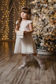 A girl in a festive light dress with a gift in her hands next to a Christmas tree, lights of garlands in the background. The concept of New Year holidays.