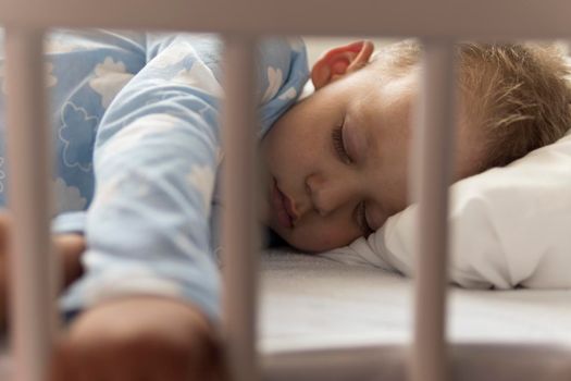 Cute cheerful little 2-3 years preschool baby boy kid sleeping sweetly in white crib during lunch rest time in blue pajama with pillow at home. Childhood, leisure, comfort, medicine, health concept.