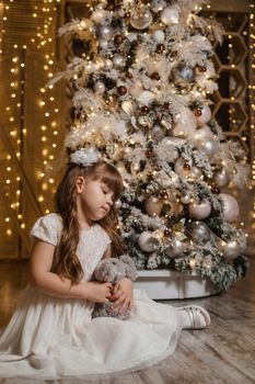 A girl in a festive light dress next to a Christmas tree, lights of garlands in the background. The concept of New Year holidays.