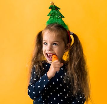 Portrait positive cheerful smiling happy little schoolgirl girl Christmas tree decoration polka dot dress biting eat orange carrots on orange background. New year, holiday, celebration, winter concept.