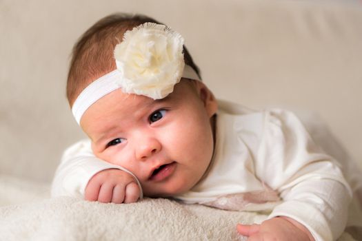 Childhood, motherhood, emotions, fashion concepts - Cute smiling happy chubby baby close up little girl in beautiful white dress flower headband on head lying on soft warm bed on her tummy at home.