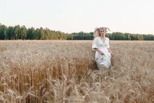 A blonde woman in a long white dress walks in a wheat field. The concept of a wedding and walking in nature.