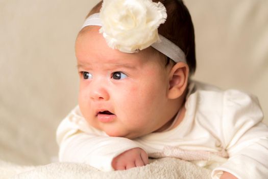 Childhood, motherhood, emotions, fashion concept - worried sad crying chubby baby with open mouth close up little girl in beautiful white dress floral headband lying on soft warm bed on tummy at home.