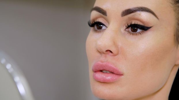 close-up, a beautiful, young woman admires her reflection in the mirror, examines her plump, sexy lips after procedure for increasing the volume of lips, correction of the shape of lips. High quality photo