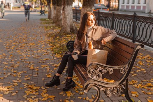 A stylish brunette woman walks around the autumn city. The brunette is sitting on a bench outside with a glass of coffee and a phone, holding a craft bag with a baguette.