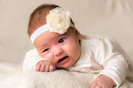 Childhood, motherhood, emotions, fashion concept - worried sad crying chubby baby with open mouth close up little girl in beautiful white dress floral headband lying on soft warm bed on tummy at home.