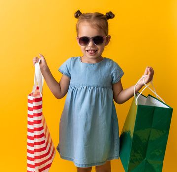 Portrait Beautiful Happy Little Preschool Girl In Sunglasses Smiling Cheerful Holding Cardboard Bags Isolated On Orange Yellow Studio background. Happiness, Consumerism, Sale People shopping Concept.