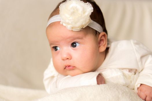 Childhood, motherhood, emotions, fashion concept - worried sad crying chubby baby with open mouth close up little girl in beautiful white dress floral headband lying on soft warm bed on tummy at home.